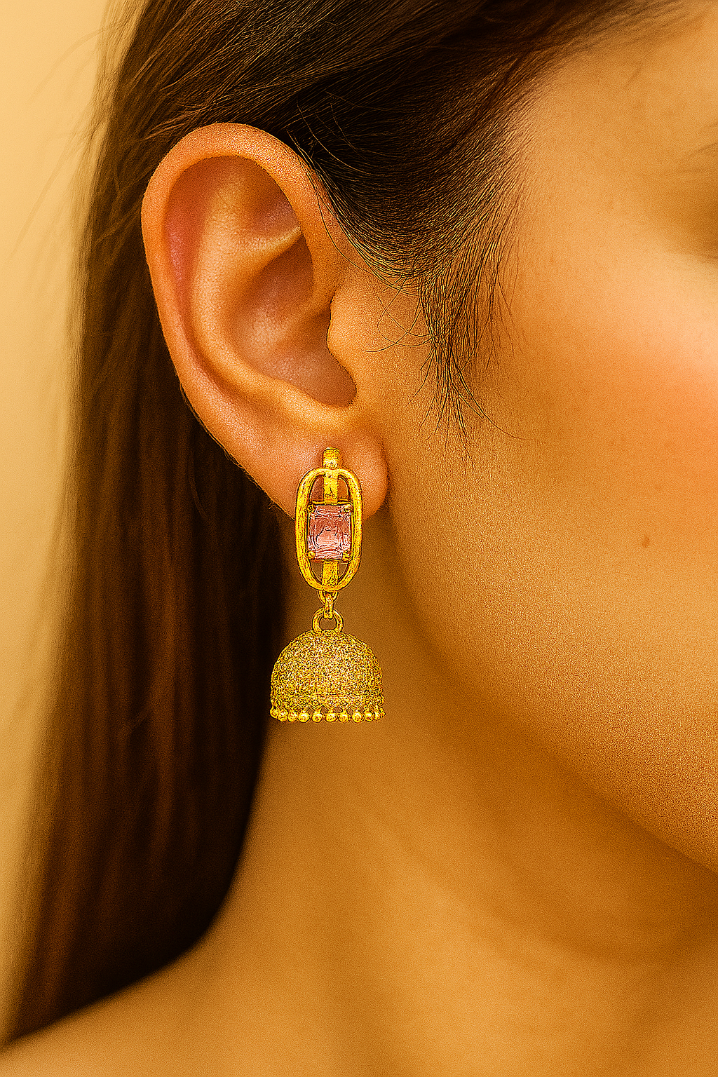 Studio-lit close-up image of a pair of gold-tone jhumka earrings featuring square-cut pink stones, hanging symmetrically against a soft beige background.